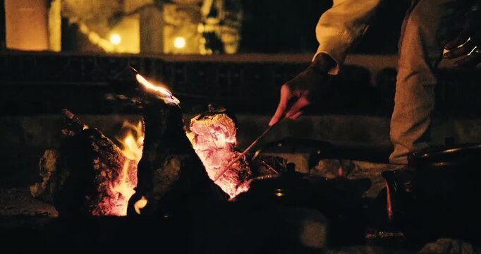 Person using a stick to tend a campfire in a rustic stone fire pit at night with glowing embers, warm flames, and a cozy outdoor atmosphere.
