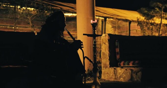 Silhouette of a person smoking shisha at a desert camp at night with warm ambient lighting, traditional seating, and a relaxed outdoor atmosphere.
