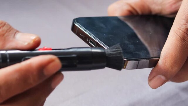 Asian man's hand cleaning a smartphone speaker grille with a small cleaning brush, also brushing around the charging port to remove dust, lint, and debris for better device maintenance and care.