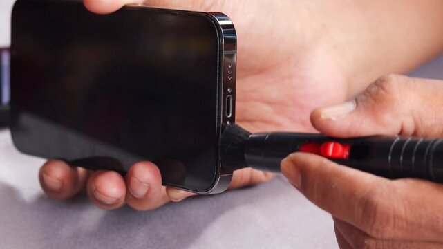 Asian man's hand cleaning a smartphone speaker grille with a small cleaning brush, also brushing around the charging port to remove dust, lint, and debris for better device maintenance and care.