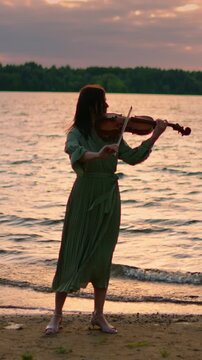 Woman in flowing green dress playing violin alone by tranquil lakeshore at golden sunset, gentle waves and distant forest framing serene musical moment