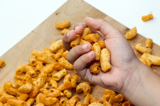 A hand holding a fistful of crispy, seasoned macaroni snacks over a wooden surface littered with the same snacks.