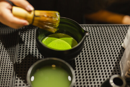 Barista lifting a traditional bamboo whisk from a black ceramic bowl of freshly whisked, frothy green matcha tea.