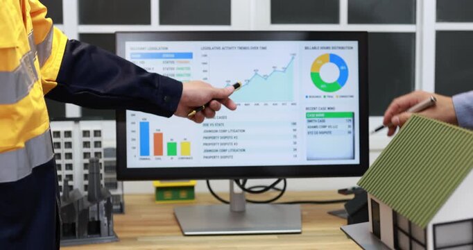 Construction workers review building project data on a computer dashboard beside a house model in a modern office.