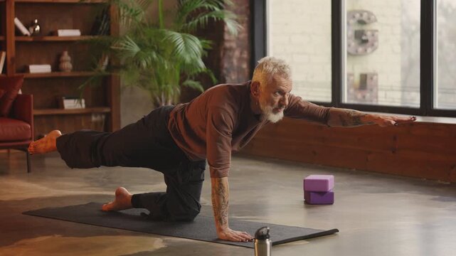 Slim silver-haired man practices tricky kitty yoga pose at home, active hobby