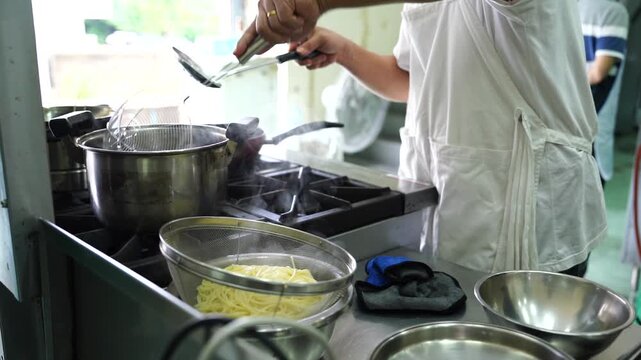 The chef is using a pot of boiling water on the gas stove to steam the spaghetti and then using a strainer to scoop it out, preparing to stir-fry the spaghetti in the kitchen.