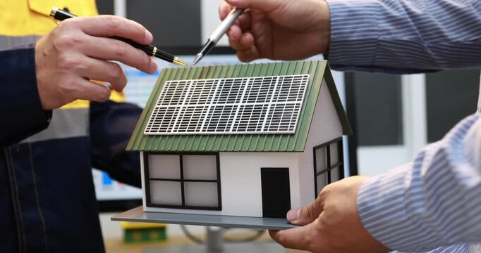 Close view of people discussing a model house with a rooftop solar panel, using pens to plan sustainable home design and renewable energy ideas.
