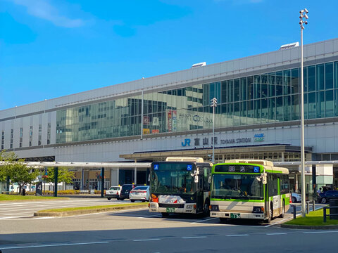 Modern exterior facade of Toyama Station with city buses parked at the terminal under a clear blue sky in Toyama Japan.