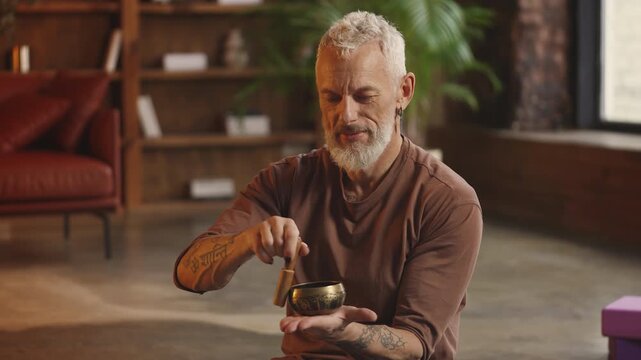 Slim man in his 50s uses Tibetan singing bowl during meditation at home