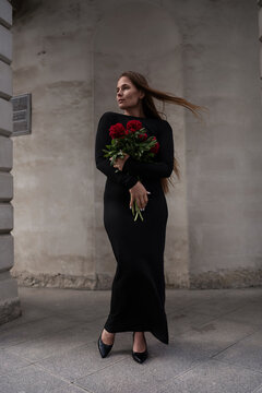 Dramatic woman posing in black dress holding vibrant red Paeonia flowers outdoors near stone architecture