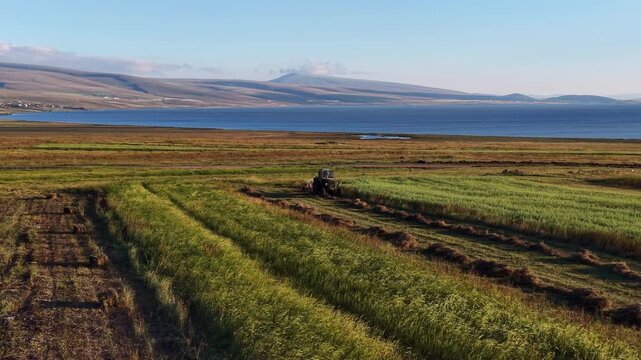 Aerial drone view of a tractor mowing hay in Georgia&rsquo;s Javakheti highlands, with Lake Paravani and expansive green pastures creating a scenic rural agricultural landscape.