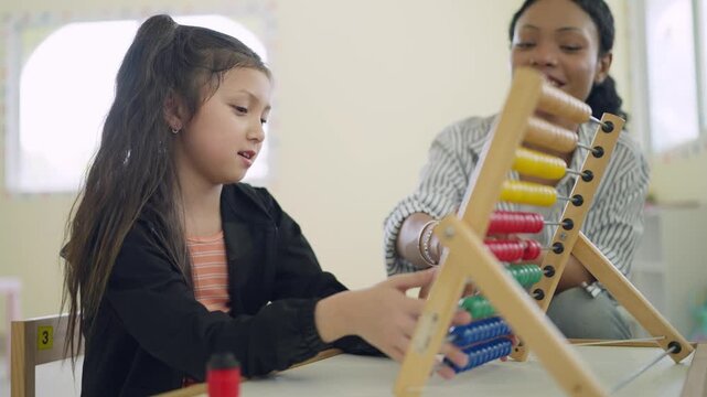 Young girl concentrates on colorful abacus with teacher beside her. Teacher smiles gently, guiding her through math learning