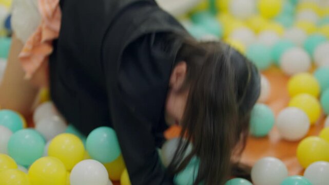Child plays in colorful ball pit with doll. Bright yellow surroundings add cheerful energy. Girl smiles, shows affection for her toy