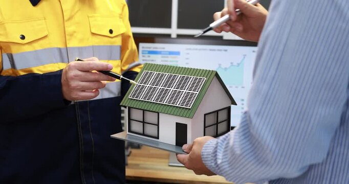 Engineer and homeowner discuss a model house with rooftop solar panels in front of an energy monitoring screen, clean power planning.