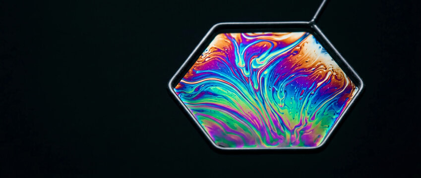 Abstract macro shot of iridescent soap film swirls inside a geometric hexagon frame; vibrant rainbow thin-film interference patterns on a dark background for psychedelic visual art.