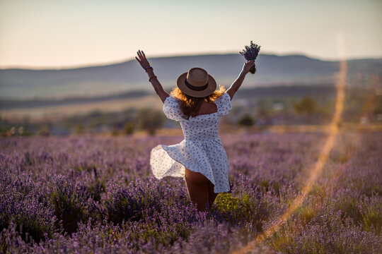 Woman lavender field, happy girl raising hands holding flowers during beautiful summer sunset.