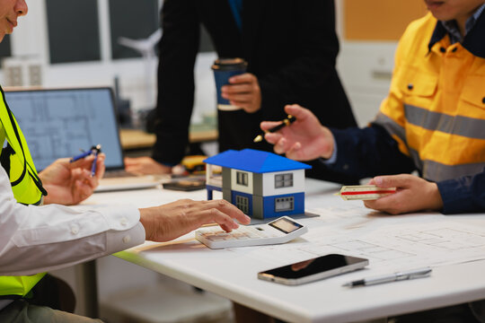 Construction team reviews house plans around a small model home, discussing building design, materials, and project details in an office.