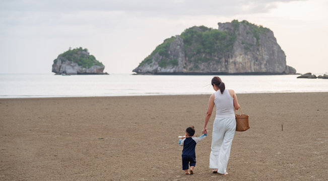 Asian mother holding hands with toddler son walking on sandy beach together.