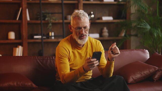 Fashionable middle-aged man scrolls social media on phone while resting on sofa