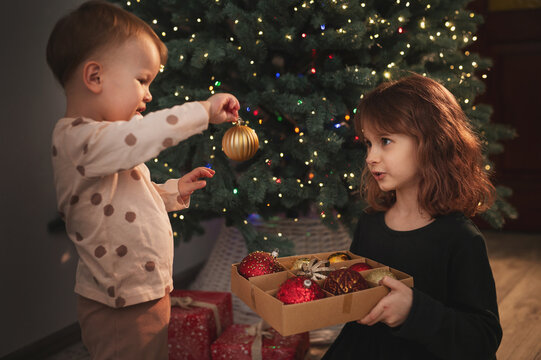 Two little girls, an older and younger sister, decorate the New Year's tree with baubles. Children are getting ready for Christmas. Christmas Eve. A magical, festive, fairytale night.