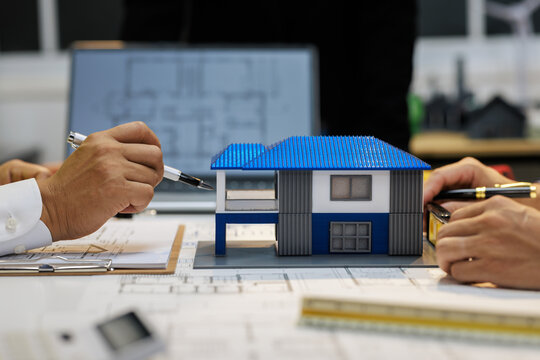 Architects review blueprints beside a small modern house model, discussing residential design and construction plans in an office.