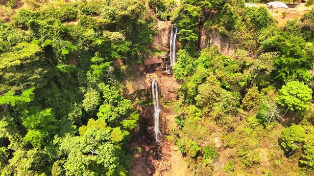 Drone pulls back and tilts upward to reveal two tall cascades of Manchewe Falls surrounded by lush green forest and dramatic cliffs in Malawi.