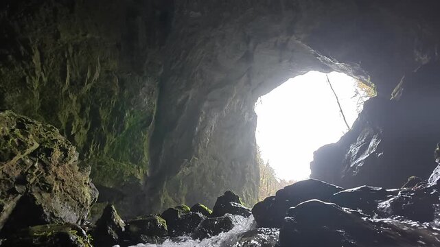 Bright sunlight illuminating the entrance of a dark cave while a river flows over rocks. The stream emerges from the underground cavern
