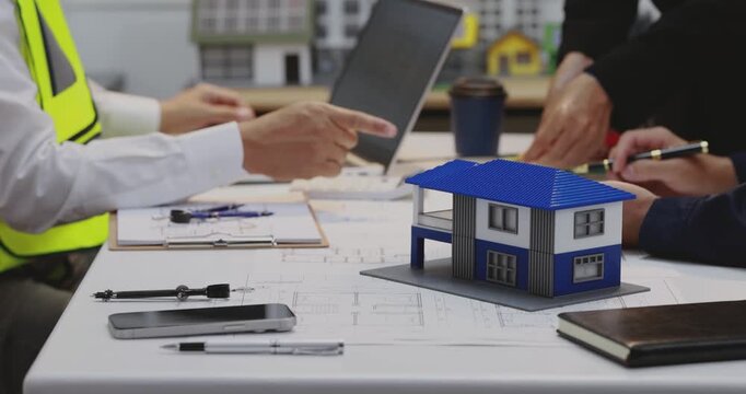 Architects and engineers discuss house plans at a desk with a blue roof model, laptop, tools and documents in a modern office.
