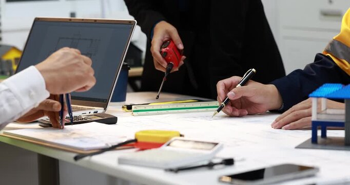 Architects and engineers review building plans at a desk with a laptop, ruler, measuring tape, calculator and small house model.