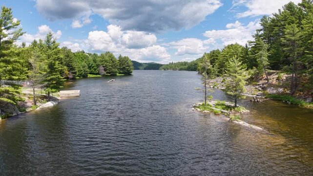 Low drone flight travels through a narrow lake channel bordered by rocky pine covered shores beneath towering summer clouds near Whitefish Falls