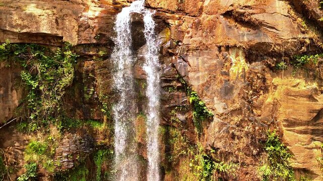 Drone descends along twin streams of Manchewe Falls in slow motion, revealing water cascading over a reddish rock cliff with patches of greenery.