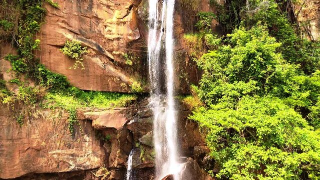 Drone rises from the base of the upper Manchewe Falls, revealing the full cascade in slow motion against a steep green escarpment in Malawi.