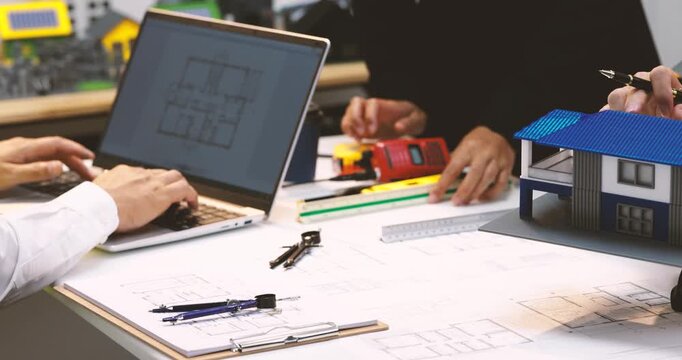 Architects review house plans with a laptop, tools, and a small modern home model on a busy design table.