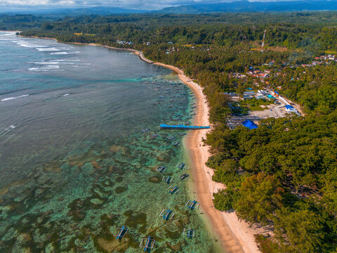 Aerial view of the coastline in Kaur Regency featuring turquoise waters, coral reefs, anchored fishing boats, a sandy beach, and lush palm trees in Kaur Regency, Bengkulu, Indonesia.
