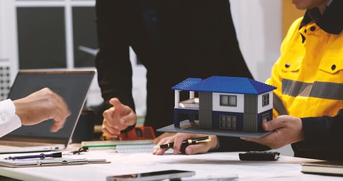 Construction team discussing a modern house model at an office table with plans, laptop and tools, showing building design, property planning and teamwork.