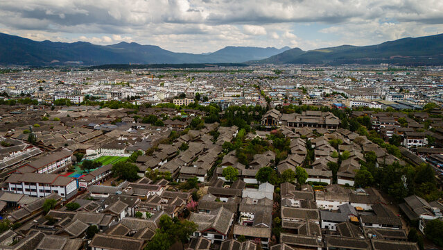 Aerial view of Lijiang Old Town with traditional grey-tiled rooftops, narrow streets, and lush trees set against distant mountains in Lijiang, Yun Nan Sheng, China.