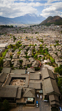Aerial view of Lijiang Old Town featuring traditional grey-tiled rooftops, lush greenery, and the majestic Jade Dragon Snow Mountain in the background in Lijiang, Yunnan, China.