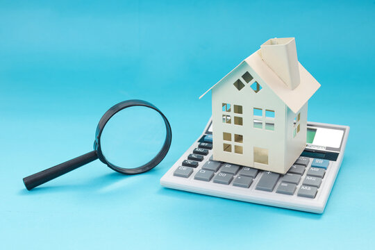 Closeup of Model House Rested on Electronic Calculator Beside Magnifying Glass Over Blue Background