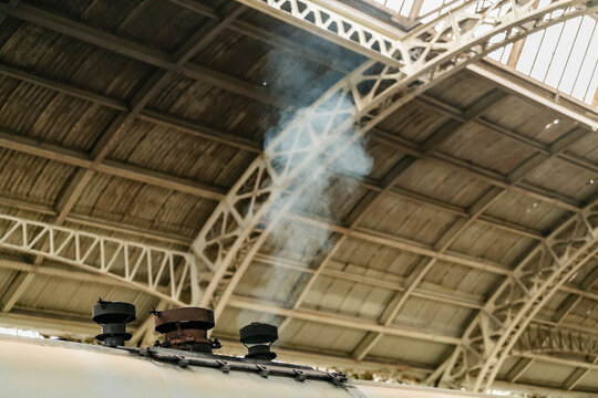 Steam locomotive chimney releasing smoke beneath a historic industrial station roof. Railway engineering, transportation heritage