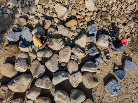 Aerial view of two workers splitting large river stones with hand tools on a sandy ground in Karanglewas, Central Java, Indonesia.