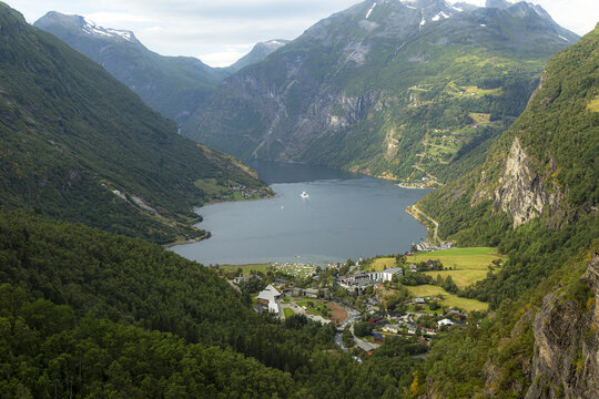 Aerial view of Geirangerfjord surrounded by steep mountains, lush green forests, and a small village nestled along the shoreline in Stranda Municipality, More og Romsdal, Norway.