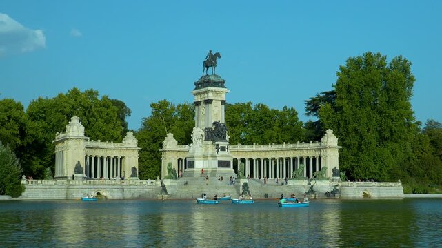 A wide shot of the iconic monument to Alfonso XII in Retiro Park, Madrid, featuring tourists in blue rowing boats on the lake under a clear sky.