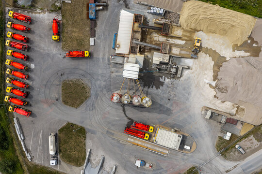 Aerial view of a cement production facility with rows of orange concrete mixer trucks parked in the yard, industrial silos, and piles of raw materials in Poland.