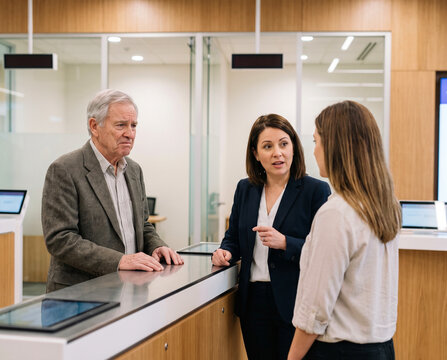 Older customer feeling ignored while bank employees speak with another client, illustrating ageism and unequal treatment in financial services.