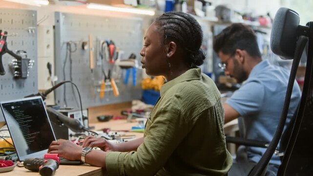 African American young adult woman typing code on laptop in robotics workshop with colleague in background. Clip is perfect for promoting engineering courses and software development services