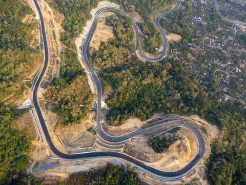 Aerial view of a winding asphalt road cutting through lush green hills and forests in Bantul, Special Region of Yogyakarta, Indonesia.
