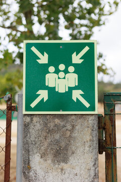 Green emergency assembly point sign with white directional arrows and group of people symbol, mounted on a weathered concrete post in an outdoor rural setting. Safety and emergency evacuation concept.