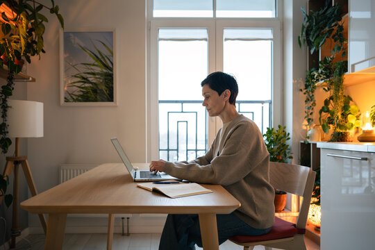 Busy mature woman working on laptop in cozy home interior with houseplants and warm lighting, self-employed freelancer female doing remote work from comfortable apartment, focused and thoughtful