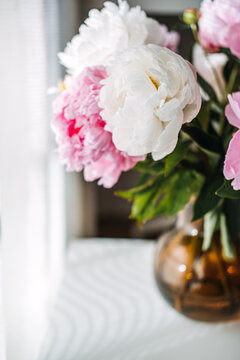 Bouquet of blooming peonies stands in glass vase on white tabletop near bright window. Modern home interior staging, minimalist apartment decor ideas, cozy living space aesthetics.