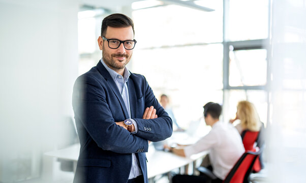 Confident mid adult businessman standing with arms crossed in front of working team